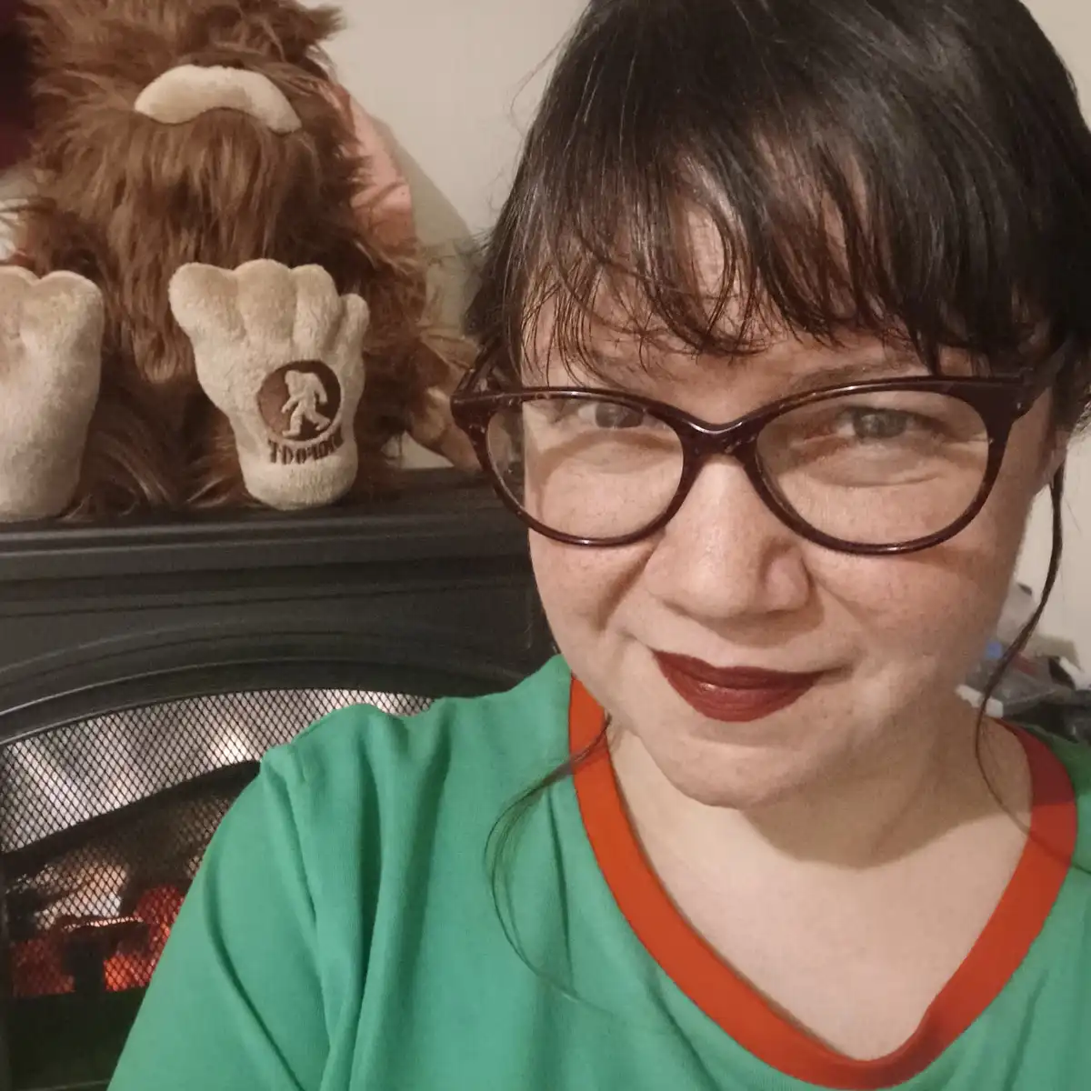 A person with dark hair and glasses smiles at the camera while wearing a green shirt with an orange collar. Behind them, on…