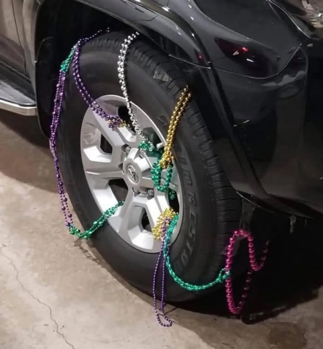 A close-up photo shows a black car's front tire wrapped in colorful Mardi Gras beads in purple, green, gold, and silver…