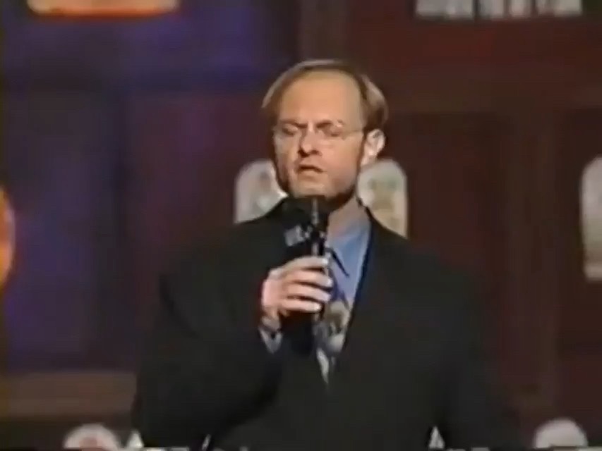 a man with glasses and a beard, wearing a dark suit and light blue shirt, holds a microphone while standing on a stage with…