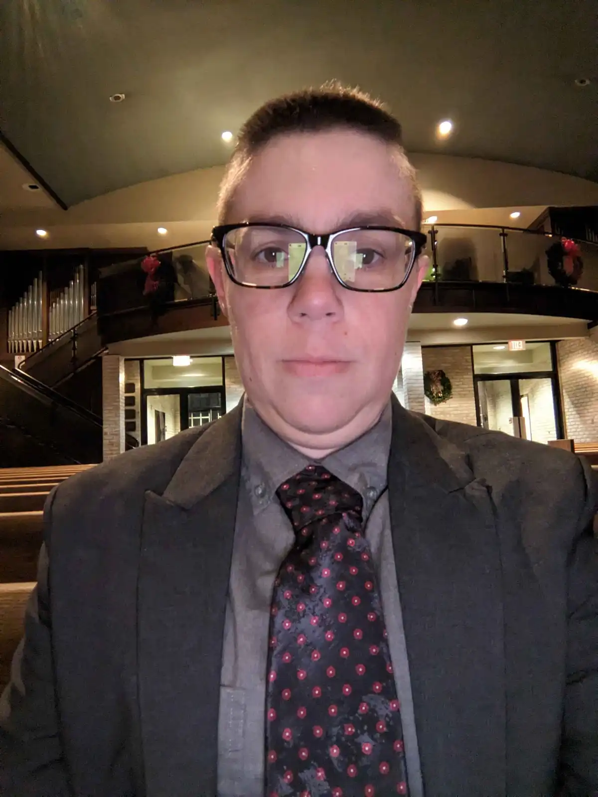 A person wearing glasses, a gray suit, and a patterned tie stands in what appears to be a church or auditorium with an organ…