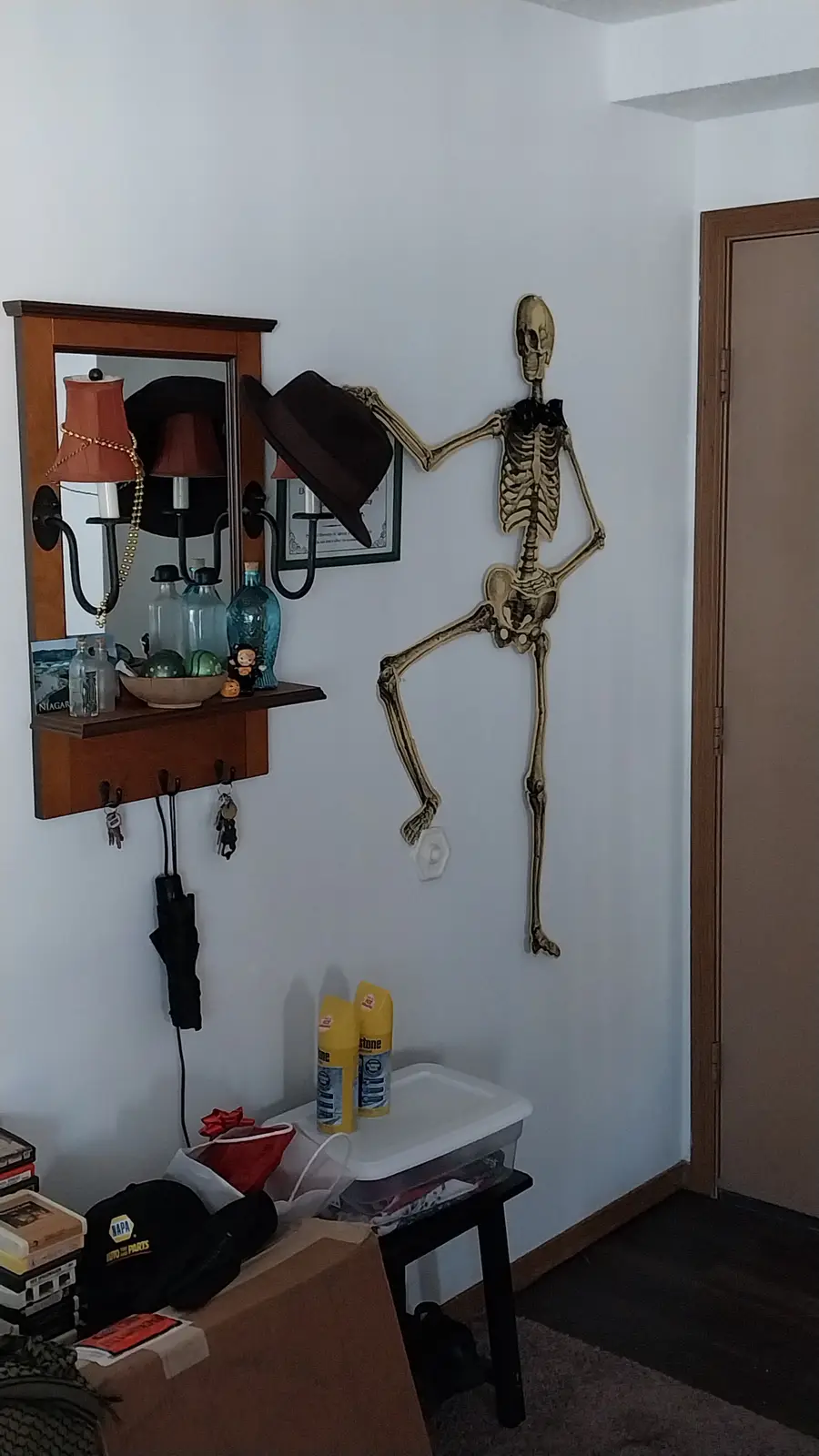 A full-sized plastic skeleton decoration is mounted on a white wall in a hallway, striking a pose with one leg kicked up and…