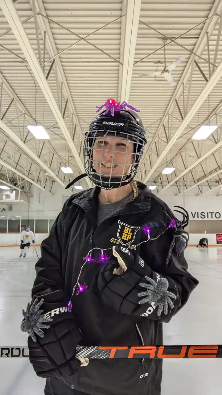 A person in hockey gear stands on an ice rink, smiling while wearing a Bauer helmet with a glowing purple spider on top…