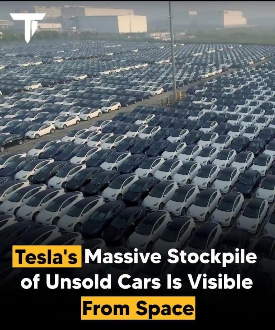 An aerial view shows a vast field filled with thousands of identical Tesla cars, mostly white and black, parked in neat…