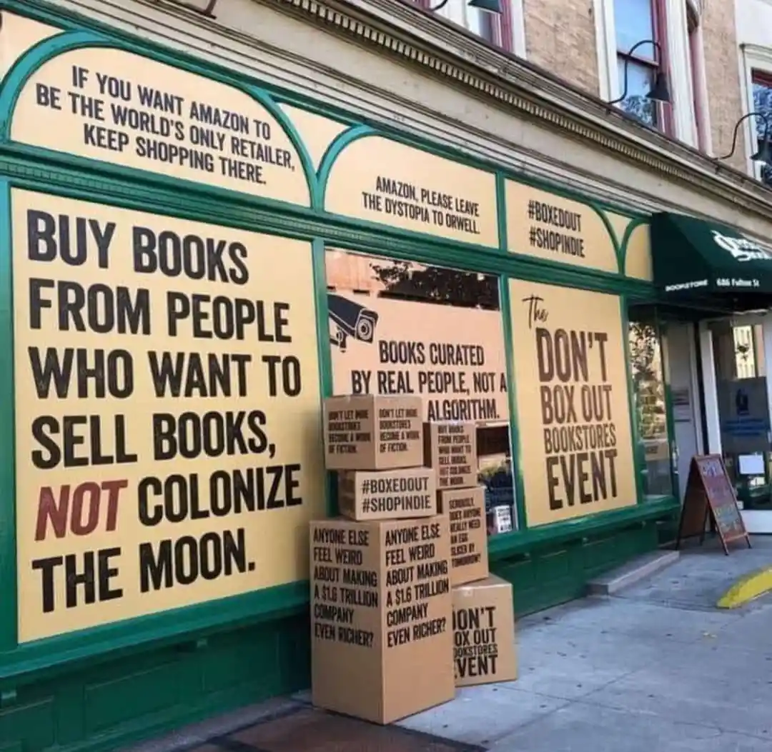 A storefront with large yellow signs and stacked cardboard boxes outside. The signs and boxes feature protest slogans…