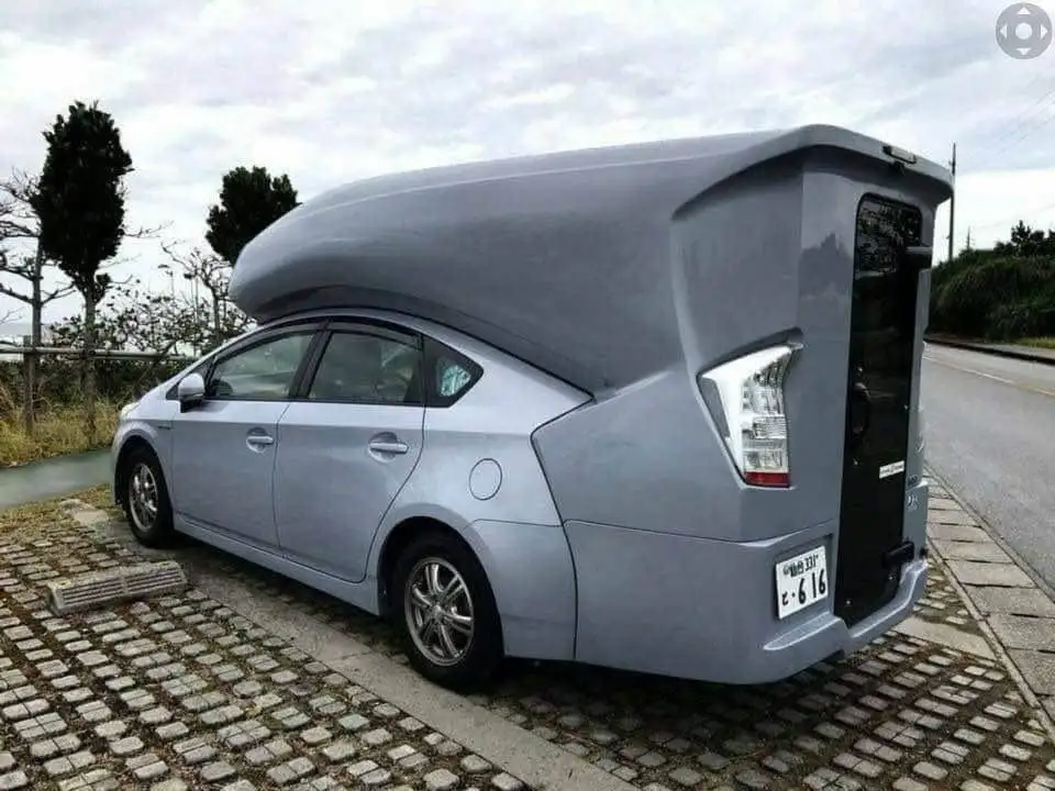 a silver toyota prius sedan is parked on a cobblestone surface with an absurdly oversized, boxy, gray camper shell attached…