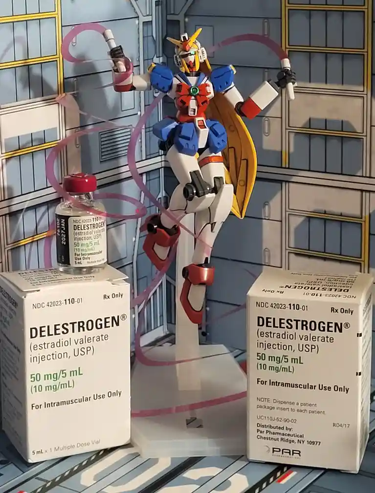A Gundam model is posed with purple tubing connected to its hands, surrounded by boxes and a vial of Delestrogen, a…