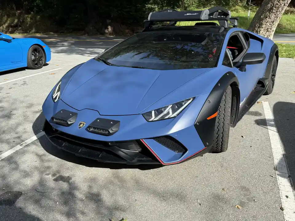 A blue Lamborghini Huracán Sterrato with a roof rack is parked in a lot, next to a blue Porsche 718 Cayman. The Lamborghini…