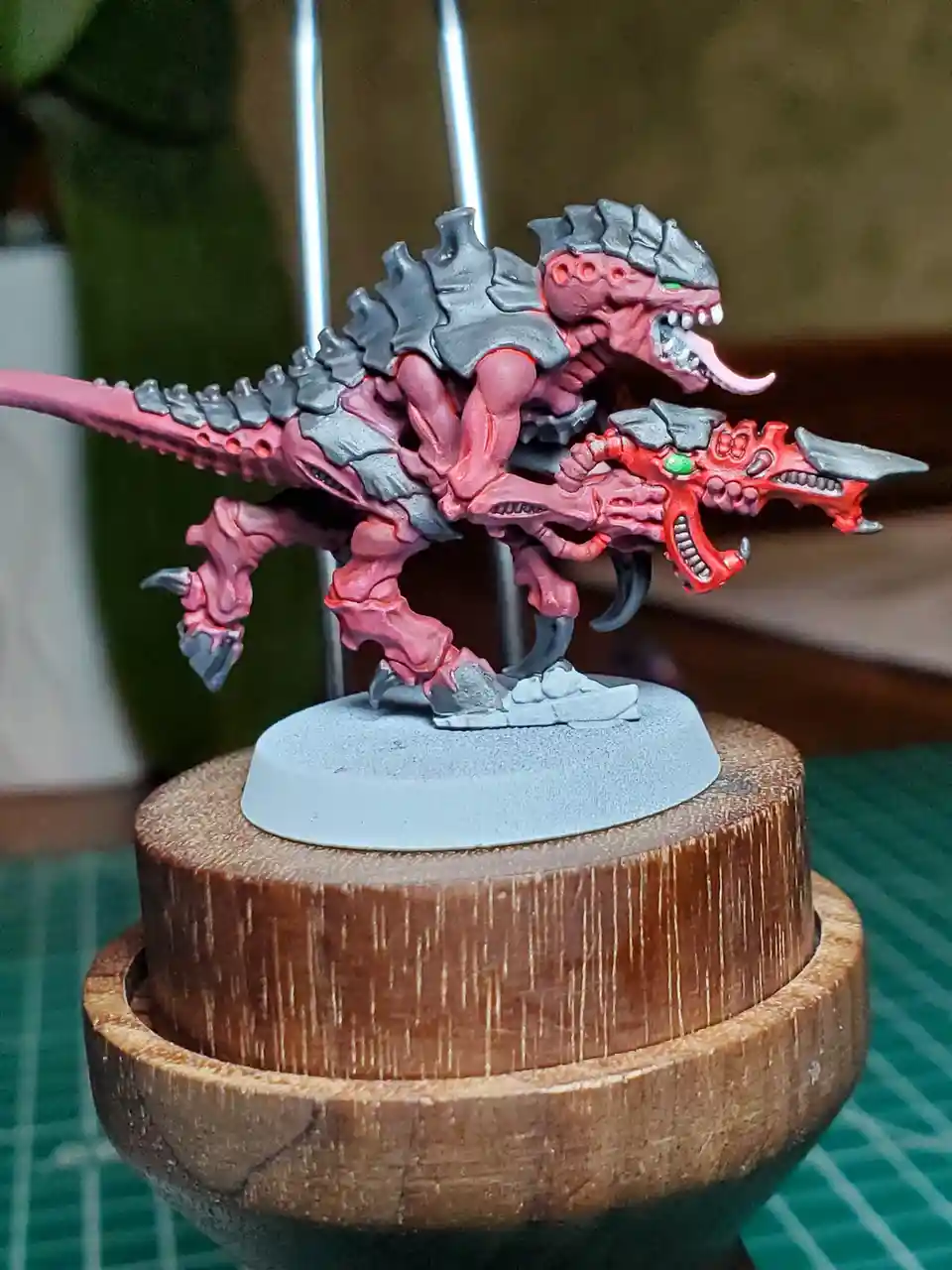 A painted miniature of a monstrous, pink and grey reptilian creature with spikes, sharp teeth, and a long tail, mounted on a…