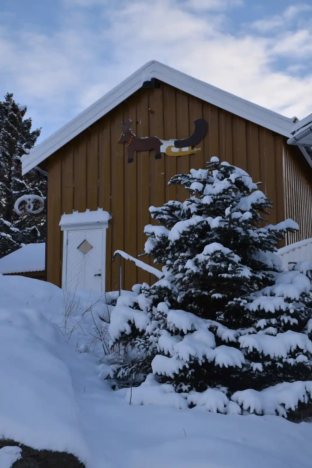 A snowy winter scene shows a wooden building with a white door and a snow-covered evergreen bush in front. A decorative…