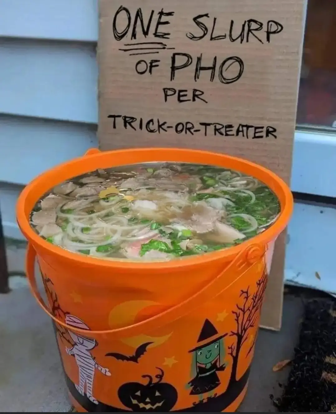 An orange Halloween-themed bucket filled with pho soup sits on a porch, with a cardboard sign behind it that reads 'ONE…