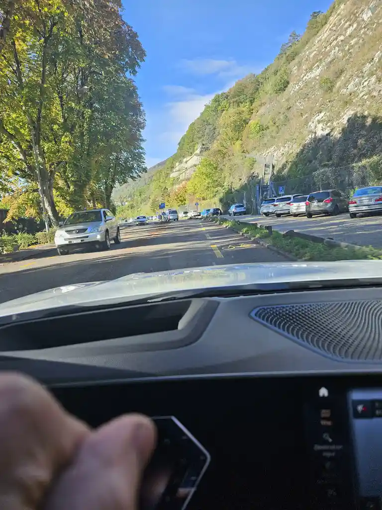 A first-person view from inside a car shows a hand holding a smartphone over the steering wheel, with a scenic road lined…