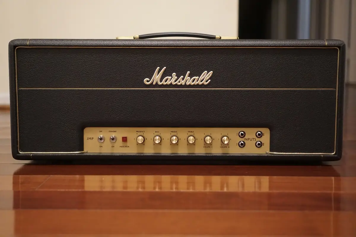 A close-up photograph of a black Marshall JMP guitar amplifier head with gold trim, sitting on a polished wooden surface…