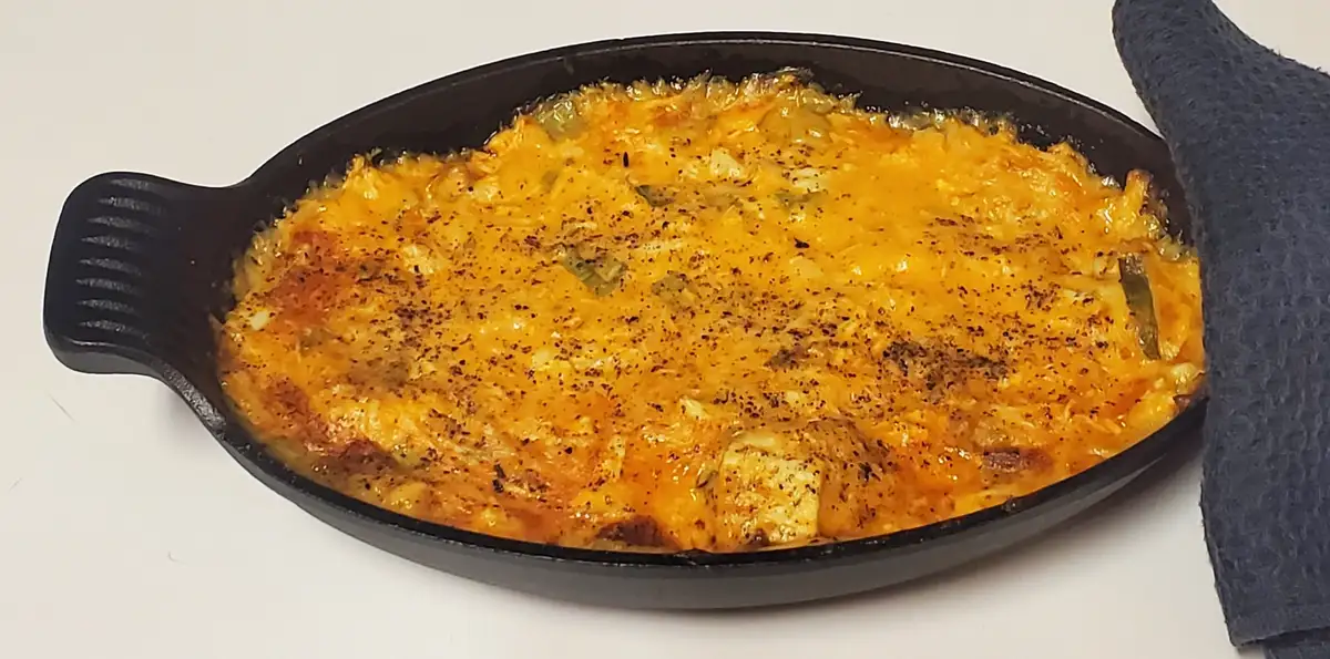 A close-up photo shows a freshly baked, cheesy casserole in a black oval cast-iron skillet, with a dark blue oven mitt…