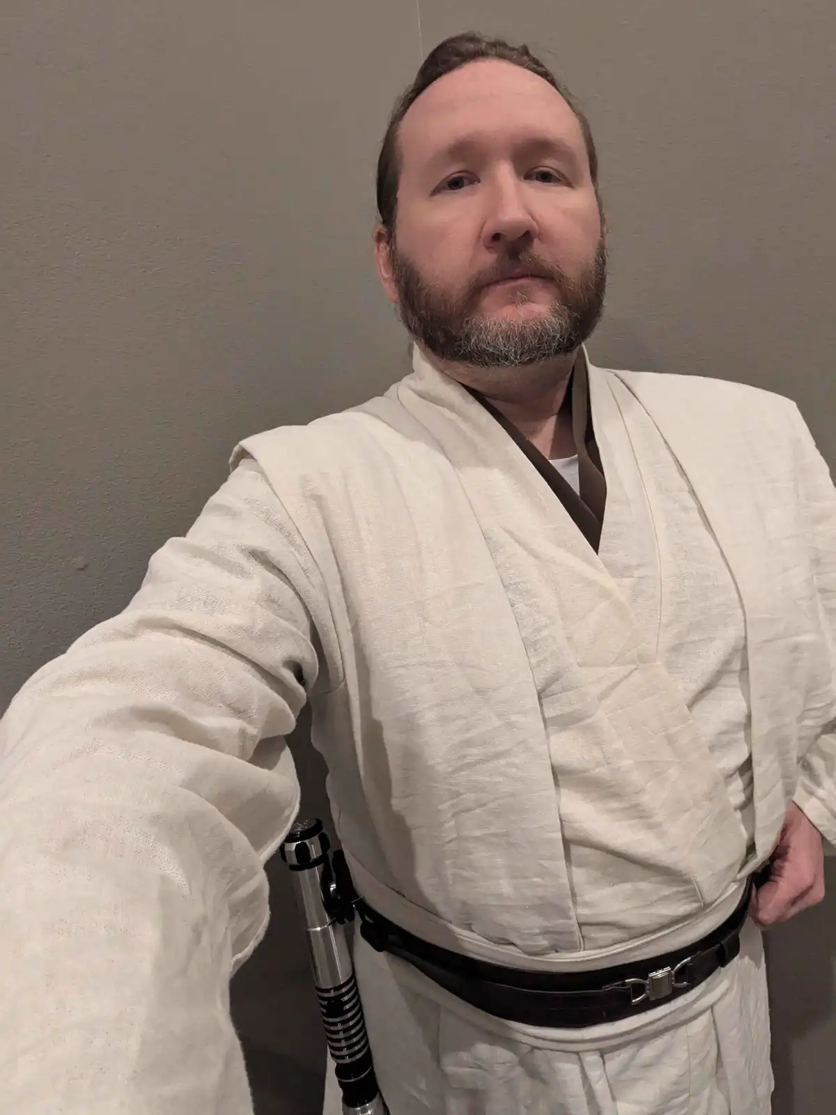 A man with a beard and gray hair takes a selfie wearing a white Jedi robe with a brown inner collar and a black belt. A…