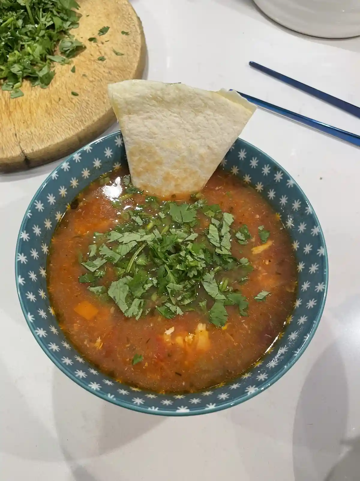 A bowl of red, chunky soup topped with chopped cilantro and a triangular tortilla chip stuck upright in it, with more…