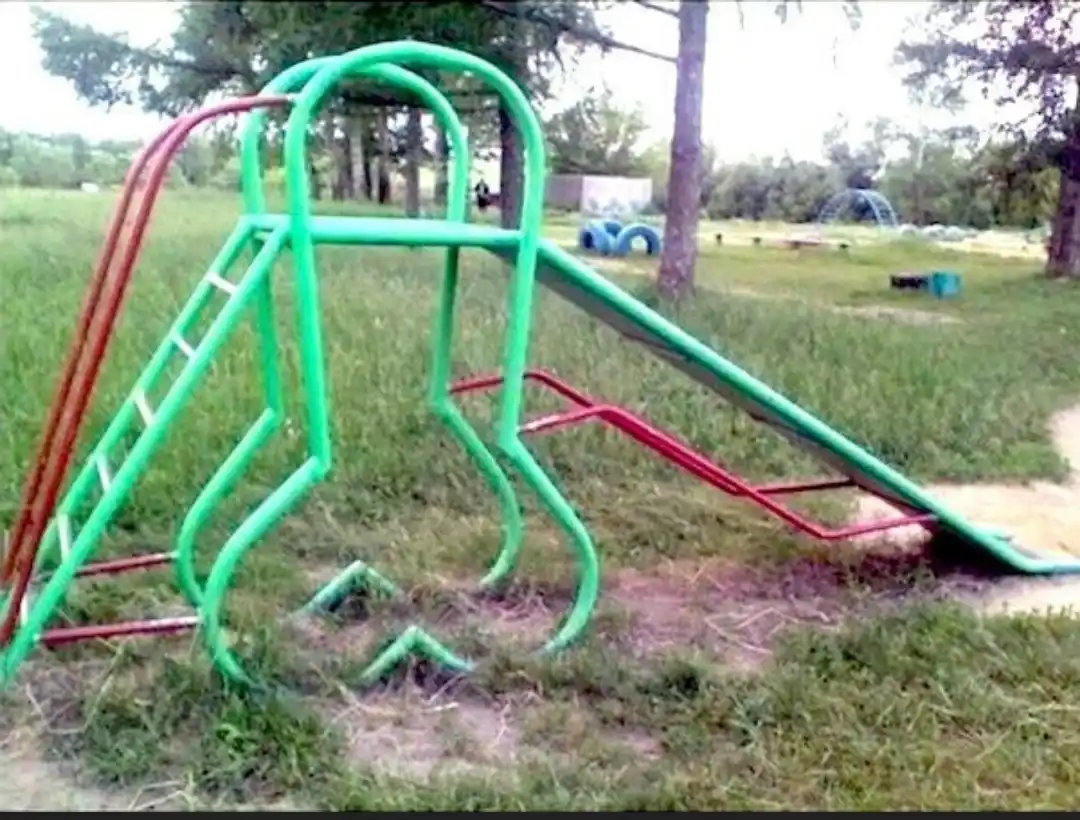 a blurry outdoor photo of a green and red metal playground slide with a ladder, set in a grassy field with trees in the…