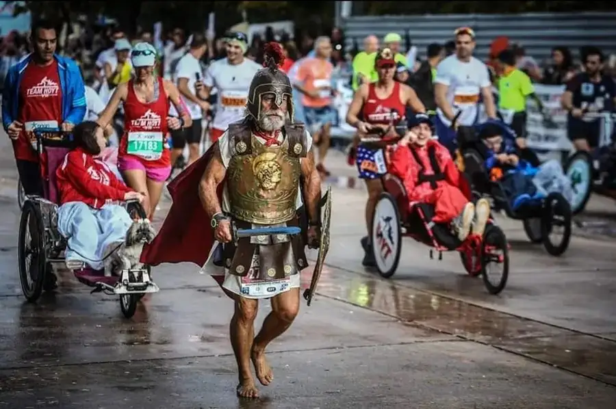 A man dressed as a Roman gladiator, barefoot and in full armor, runs barefoot in a race while others push wheelchairs with…