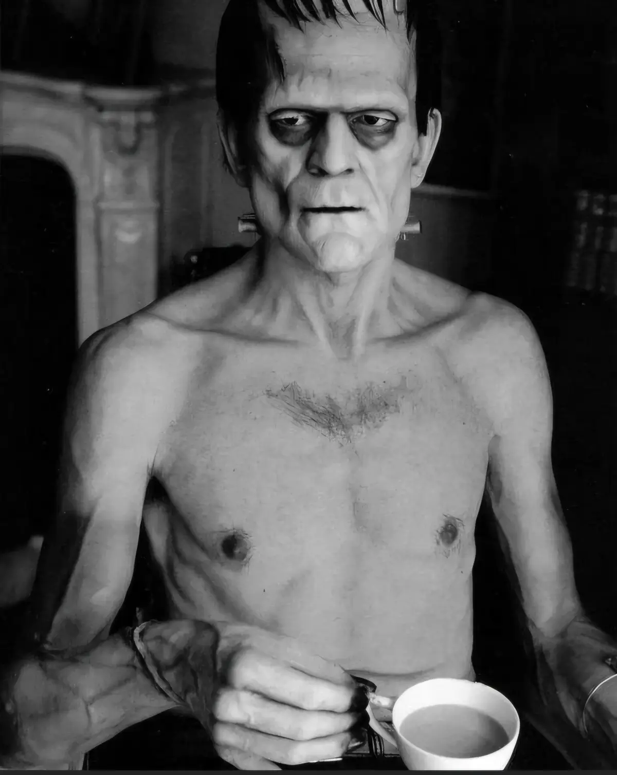 A black-and-white photo shows a shirtless, classic Frankenstein monster with bolts on his neck, holding a white coffee mug…