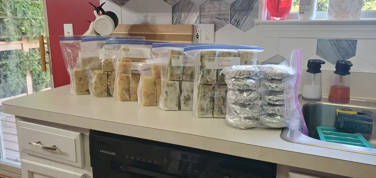 A kitchen counter displays multiple labeled zip-top bags filled with frozen, pre-portioned meals like 'Chicken Lasagna' and…