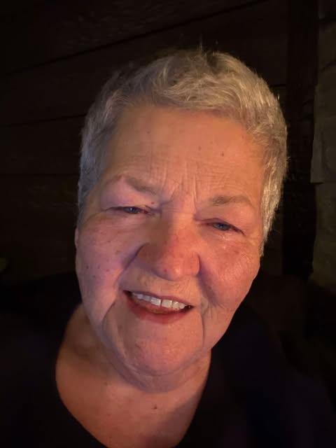 A close-up photo of an older woman with short gray hair, smiling warmly at the camera. She has visible wrinkles and…