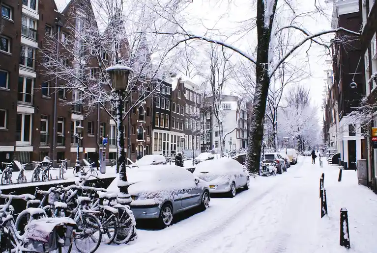 A snowy street scene in Amsterdam, featuring snow-covered cars, bicycles, and trees lining a canal-side road with brick…