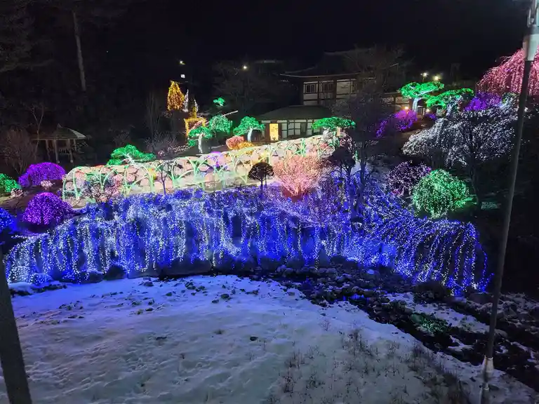 a nighttime photo of a snowy landscape featuring a rock cliff face and a small stream, both heavily decorated with colorful…
