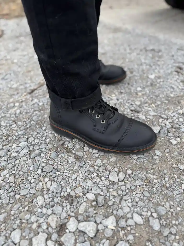 A close-up shot of a person's lower leg and foot wearing black leather boots with black laces and a brown sole, standing on…