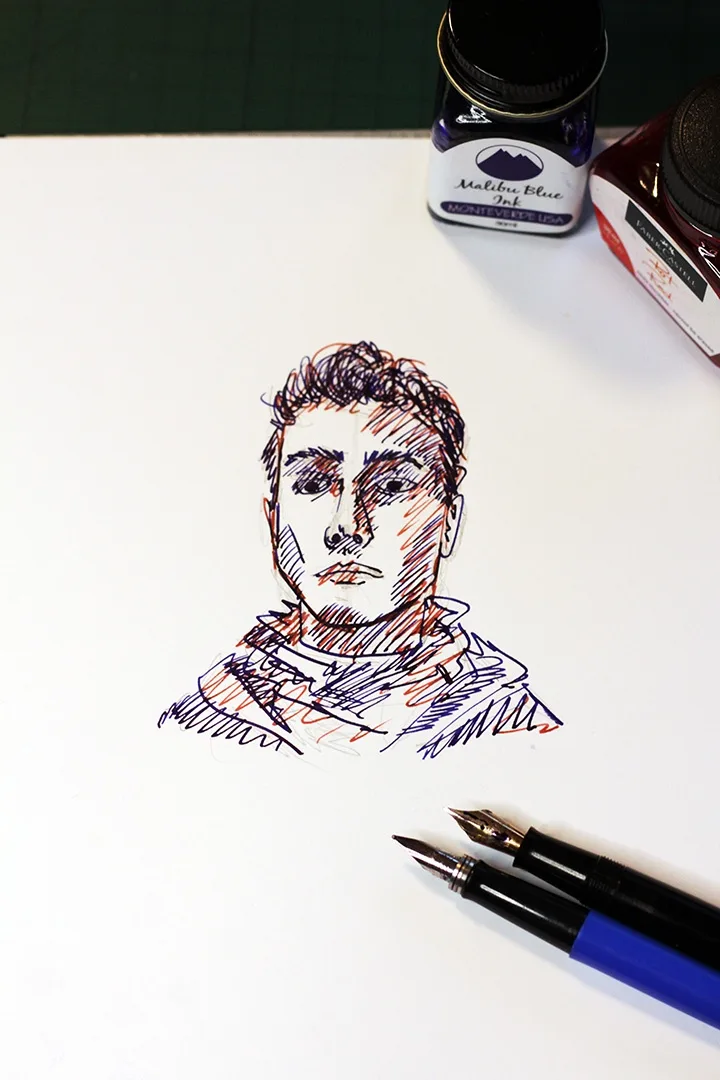 A hand-drawn portrait on white paper, rendered with blue and red ink using a fountain pen, sits beside two fountain pens and…