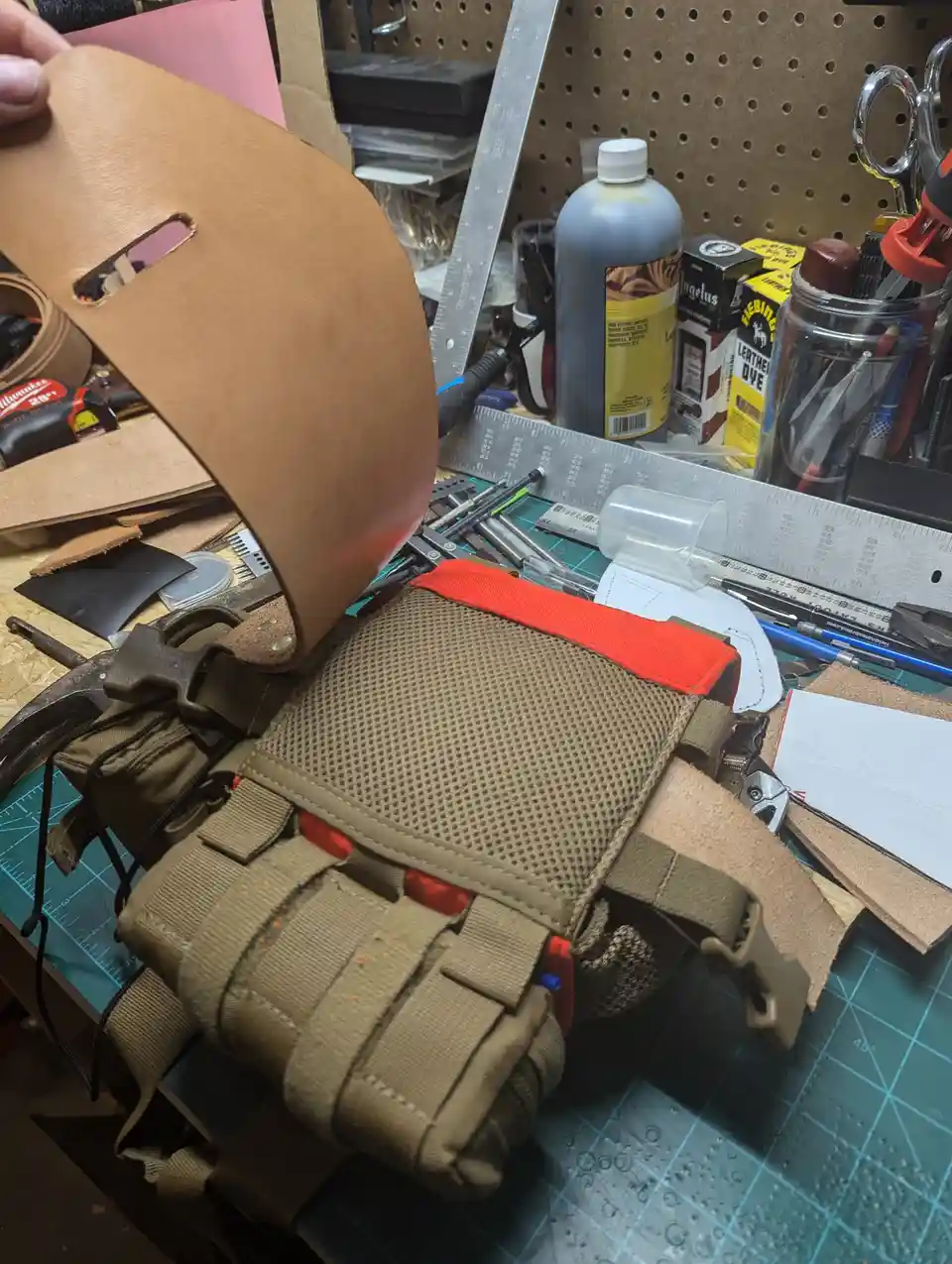 A cluttered leatherworking bench shows a person holding a tan leather piece with a cutout, next to a tan and orange…