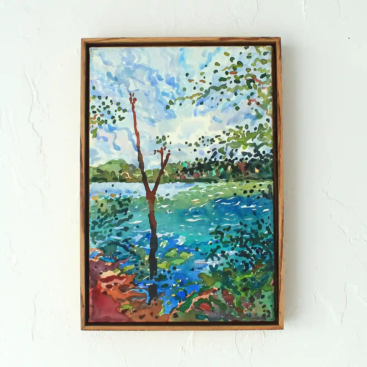 A watercolor painting in a wooden frame hangs on a white wall, depicting a stylized landscape with a prominent, bare brown…