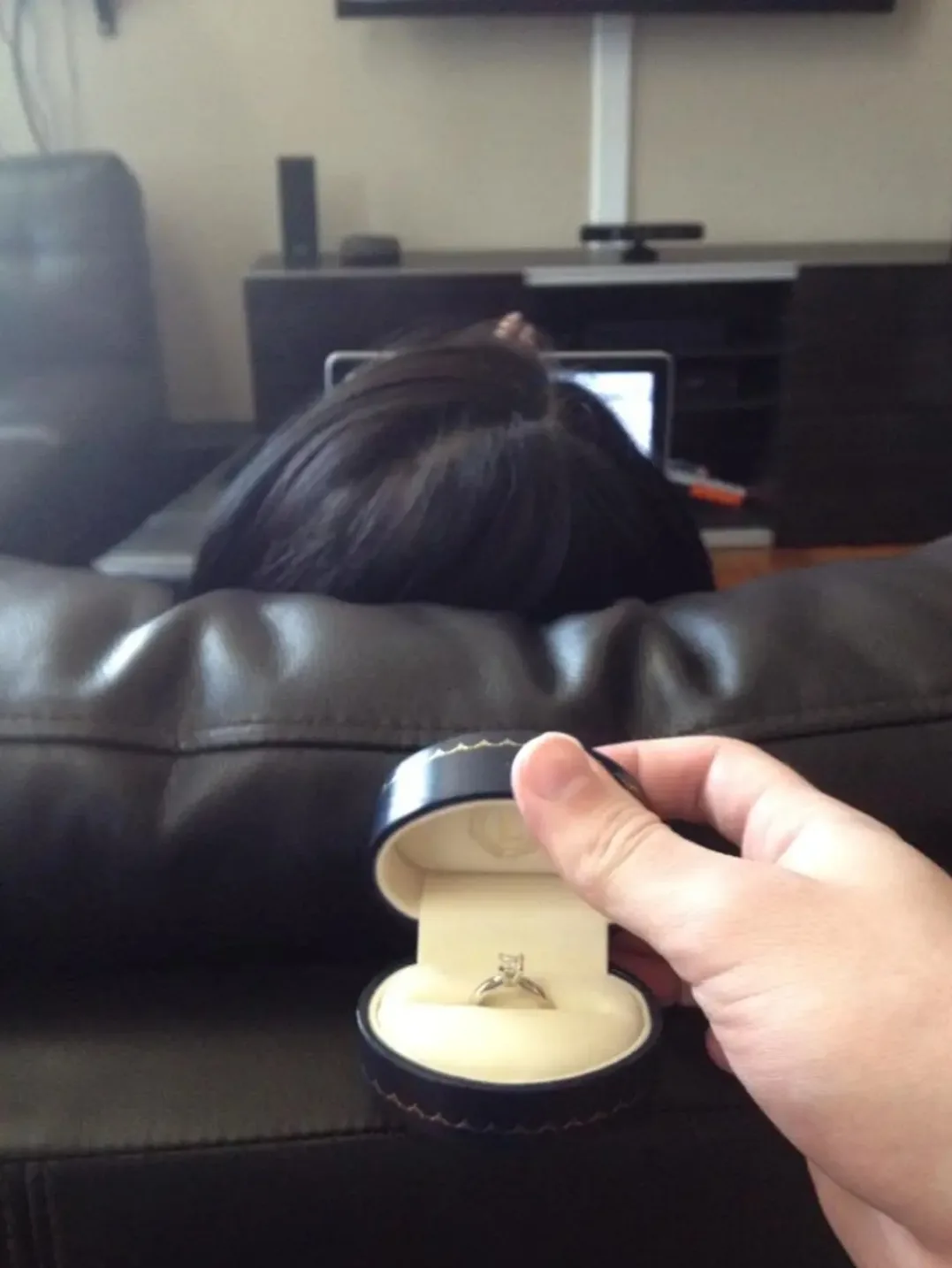 A hand holds an open black ring box with a diamond engagement ring inside, positioned in front of a person with dark hair…