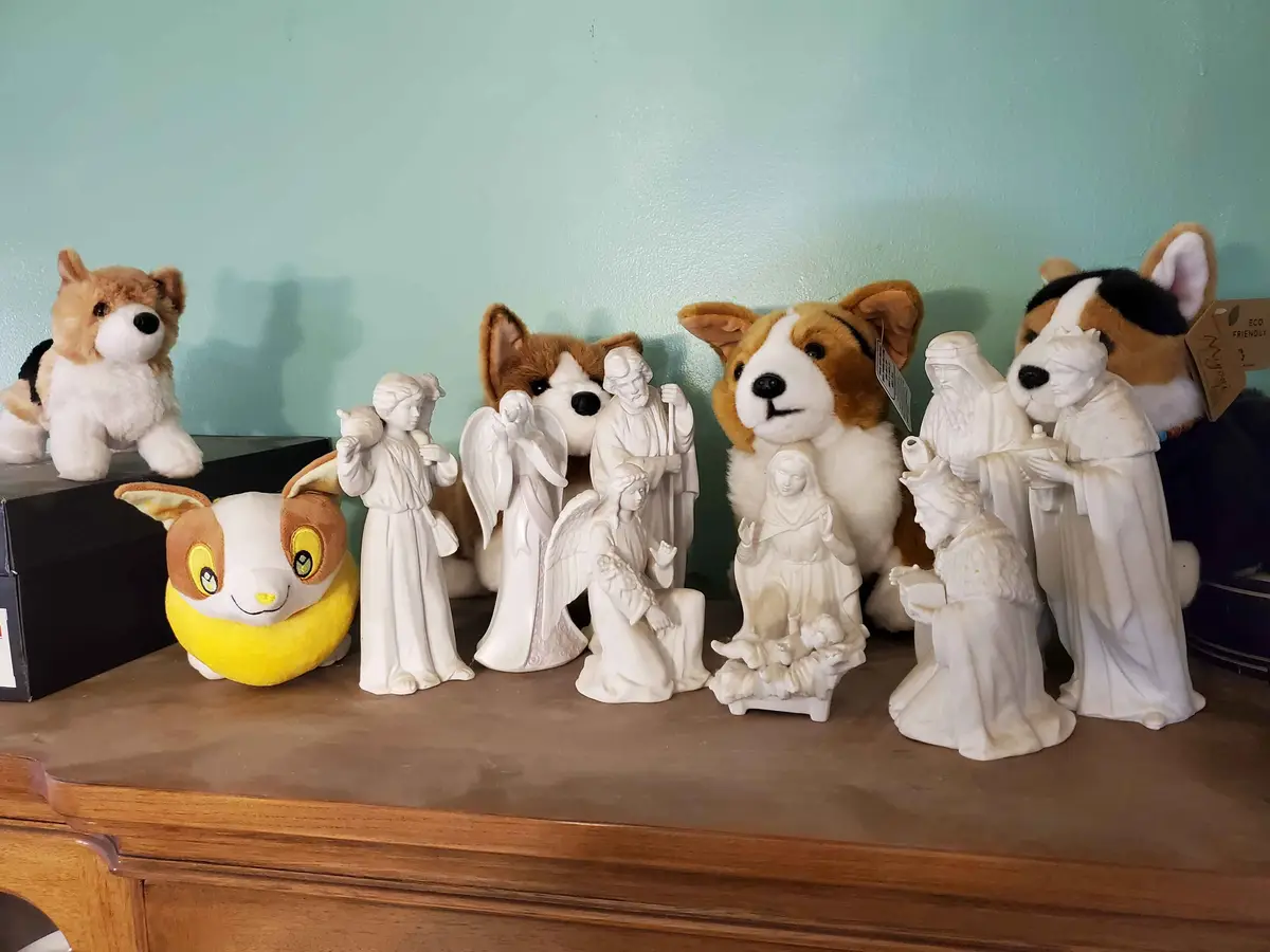 A collection of white ceramic nativity scene figurines is arranged on a wooden surface, with several plush corgi toys…
