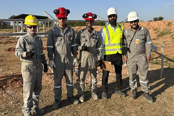 Five men in work overalls and safety gear stand outdoors at a construction site. Four wear hard hats (yellow, red, red…