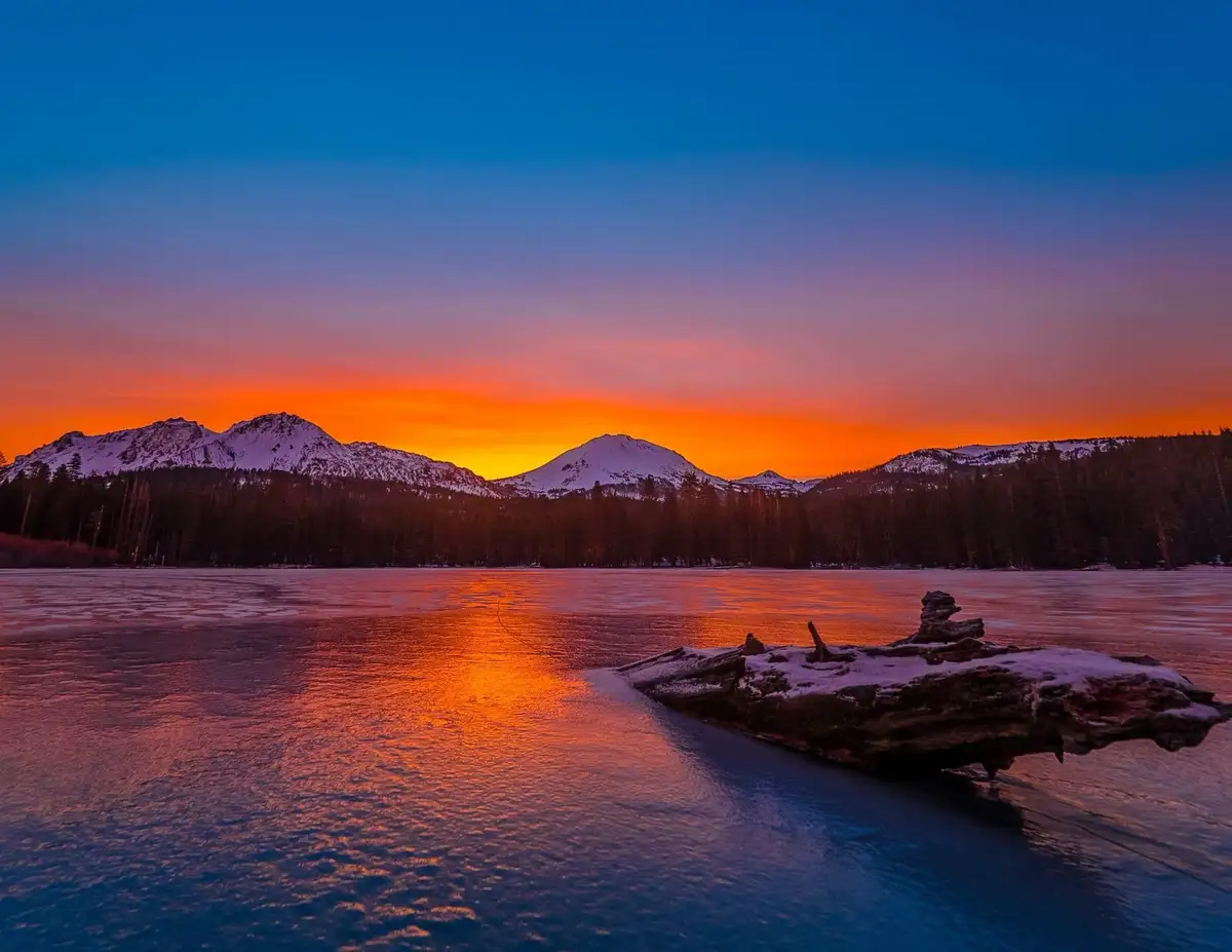 A serene winter landscape shows a frozen lake reflecting a vibrant orange and blue sunset over snow-capped mountains, with a…