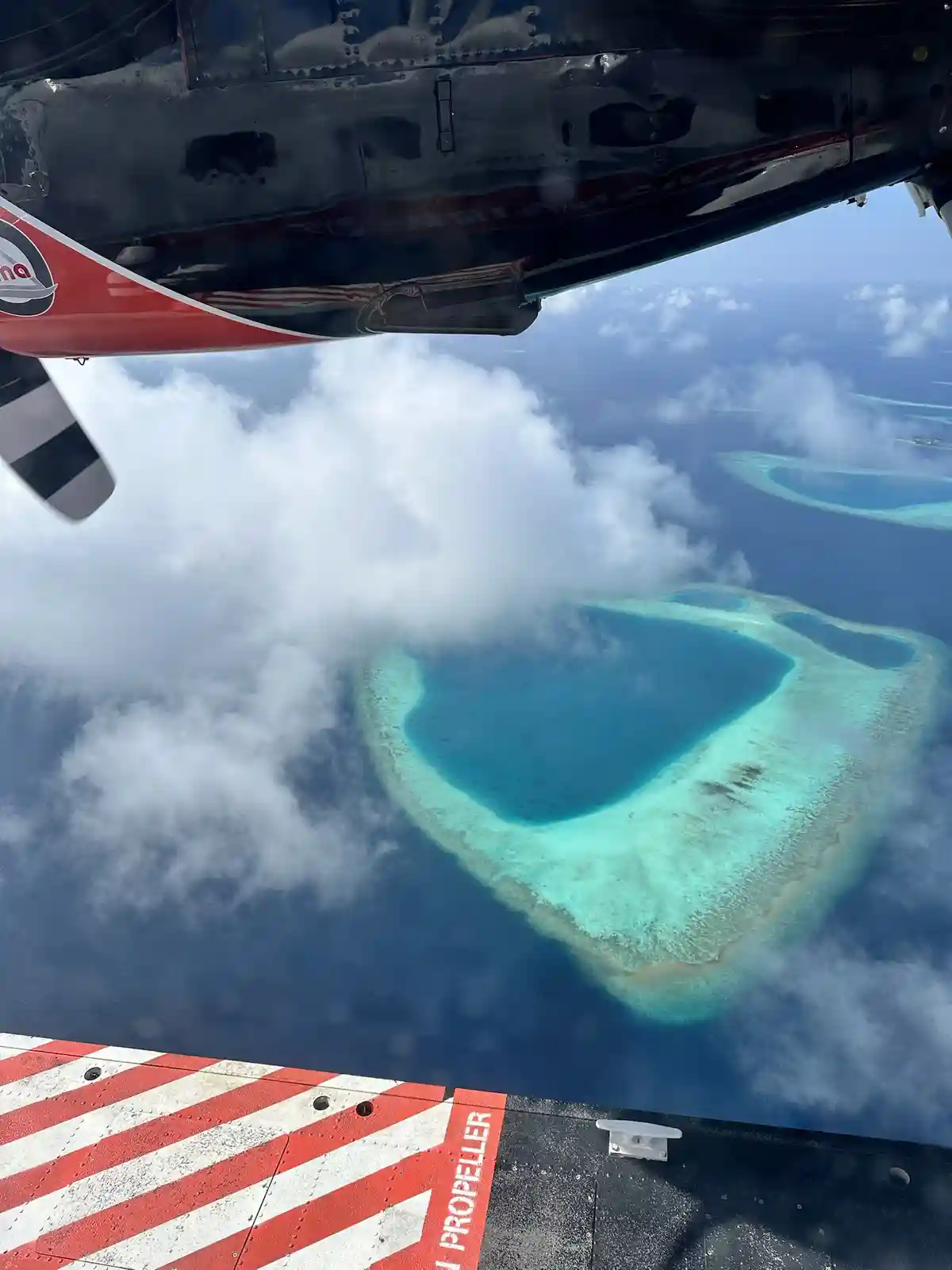 An aerial view from inside a small airplane shows the red and black wing and propeller of the aircraft, with a red and white…
