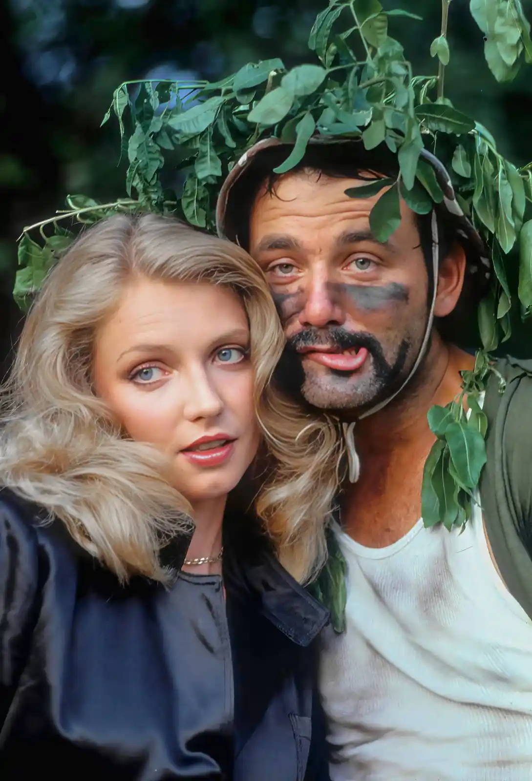 A man with a leafy crown and camouflage face paint sticks his tongue out playfully while a woman with blonde hair and a…