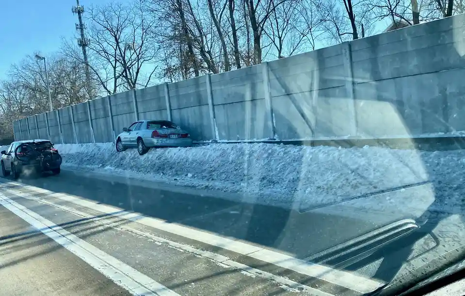 A silver sedan is parked on a snowbank next to a concrete highway barrier, appearing to be stuck or abandoned. Another car…