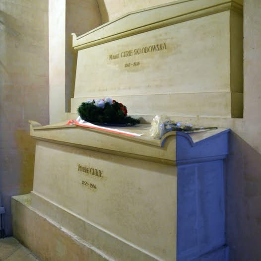 A stone tomb monument with two engraved plaques: the top reads 'Marie Curie-Skłodowska 1867-1934' and the bottom reads…