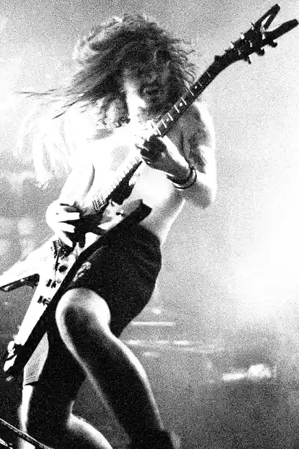 A grainy black-and-white photo captures a shirtless, long-haired rock guitarist mid-performance, his hair flying as he plays…
