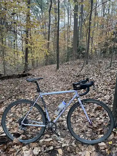 A light purple bicycle with black handlebars and a water bottle is parked on a trail covered in fallen autumn leaves. The…