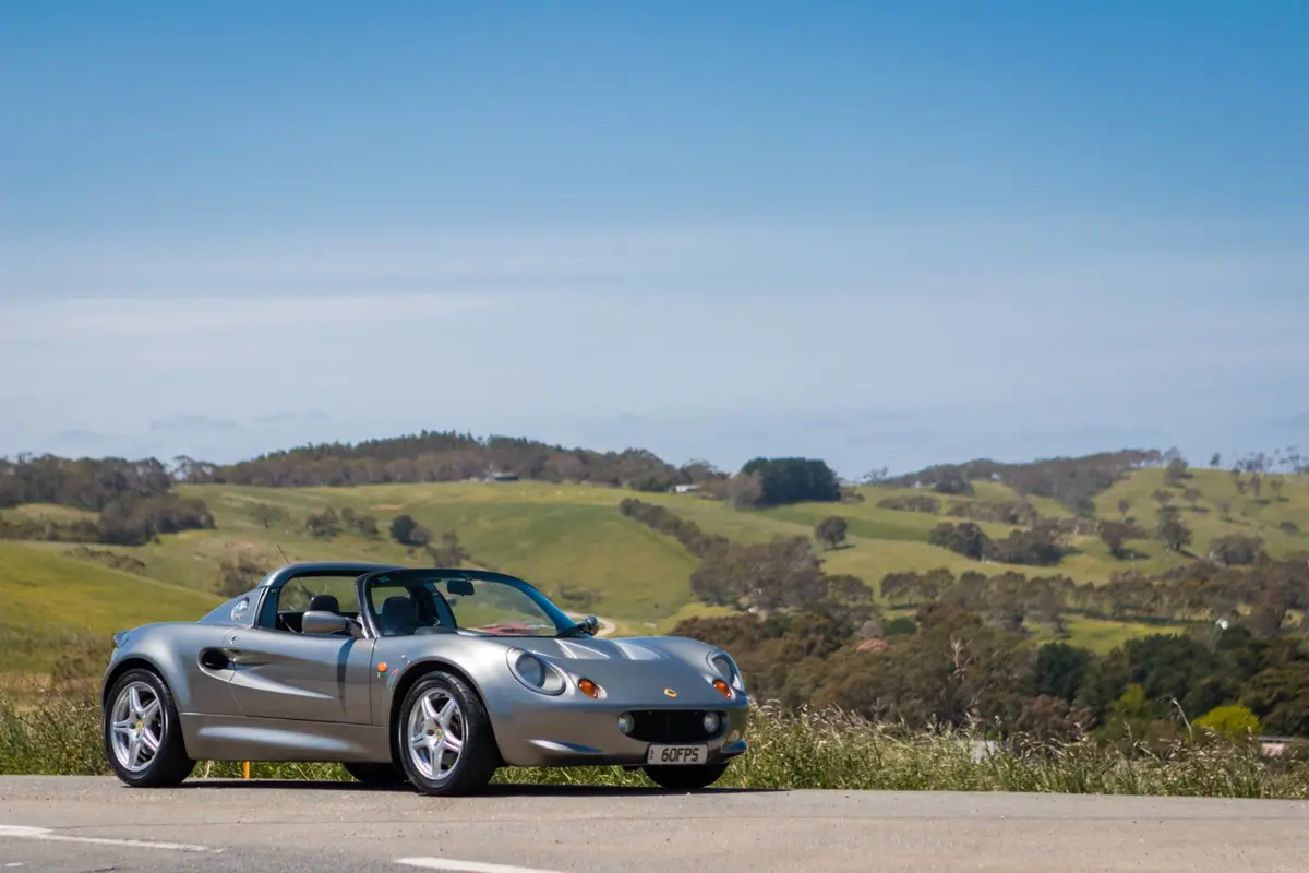A silver Lotus Elise sports car is parked on a roadside with rolling green hills and trees in the background under a clear…