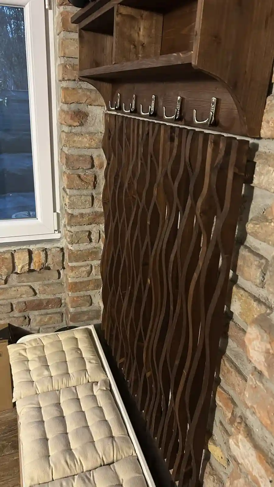 A wooden coat rack with six metal hooks is mounted on a brick wall next to a window, with a tufted bench below. The overlaid…