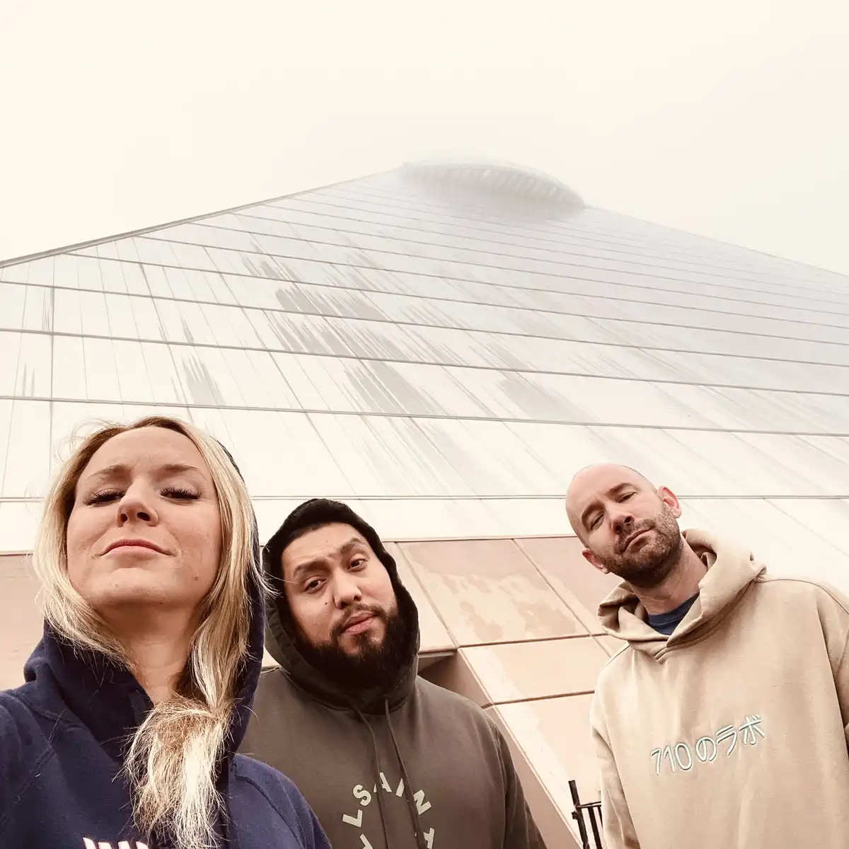 Three people stand in front of a modern, angular building partially obscured by fog, all looking upward with expressions of…