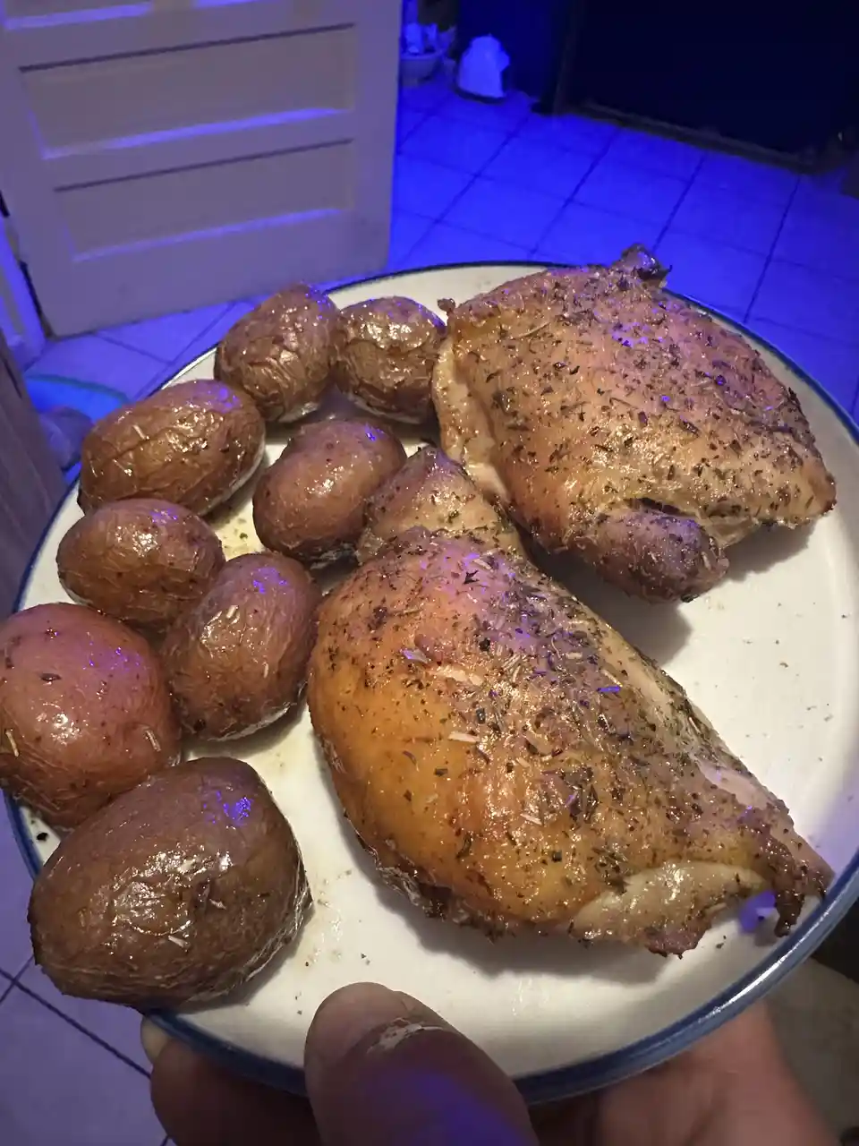 A close-up photo shows a white plate with a blue rim being held by a person's hand, containing two roasted chicken thighs…