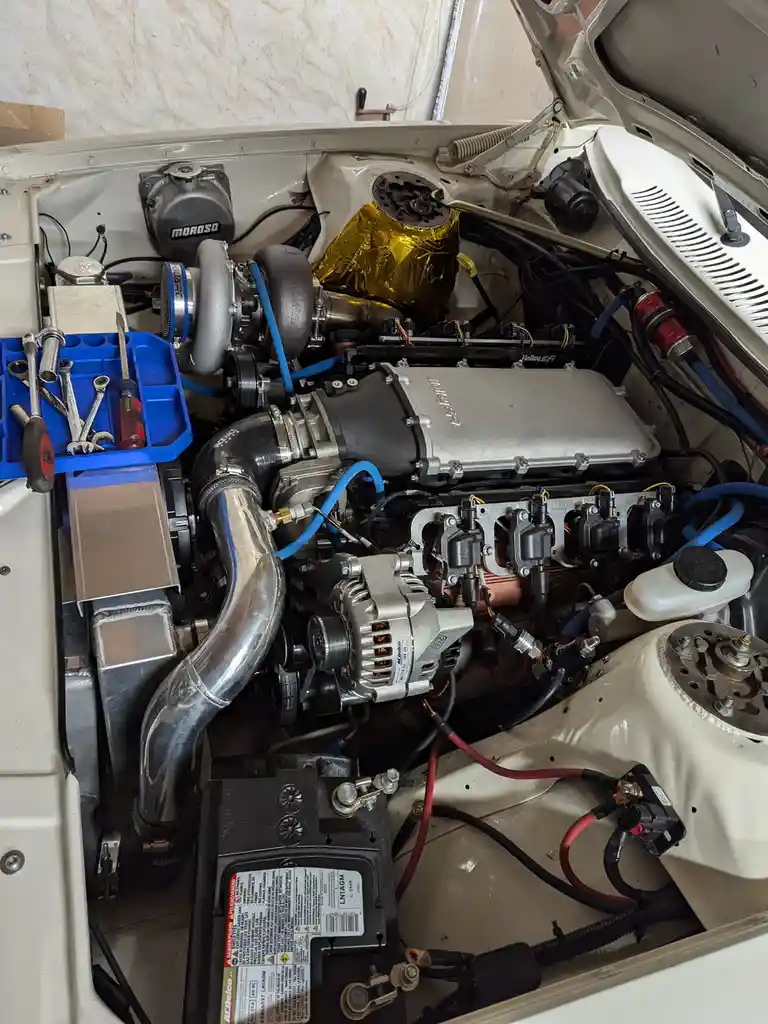 A close-up photo of a heavily modified car engine bay with a turbocharger, custom piping, and various aftermarket parts. A…