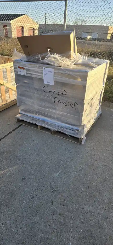 A large, heavily shrink-wrapped cardboard box sits on a wooden pallet outdoors, with handwritten text on its side reading…