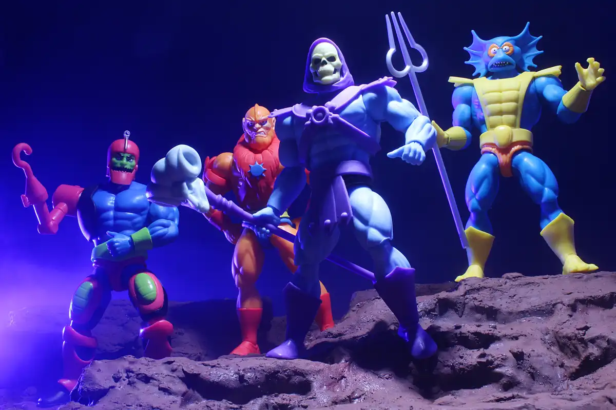Four He-Man action figures, including Skeletor with a purple hood and trident, stand on a rocky terrain under purple…