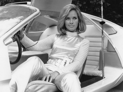 a woman in a white futuristic outfit sits in the driver's seat of a sleek, white, retro-futuristic car with the door open…