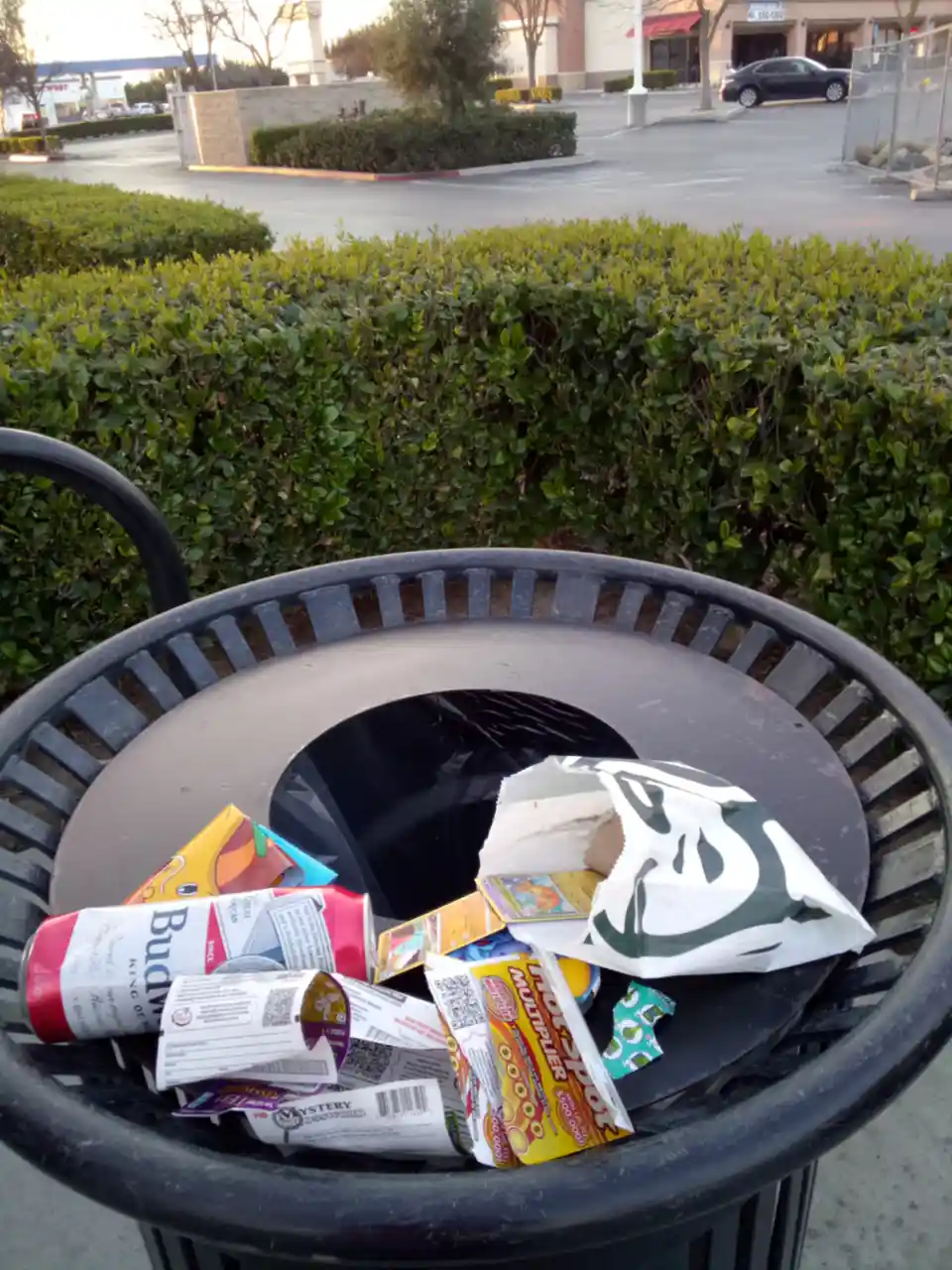 A black outdoor trash can is overflowing with discarded items including a Budweiser can, a Starbucks bag, and various candy…