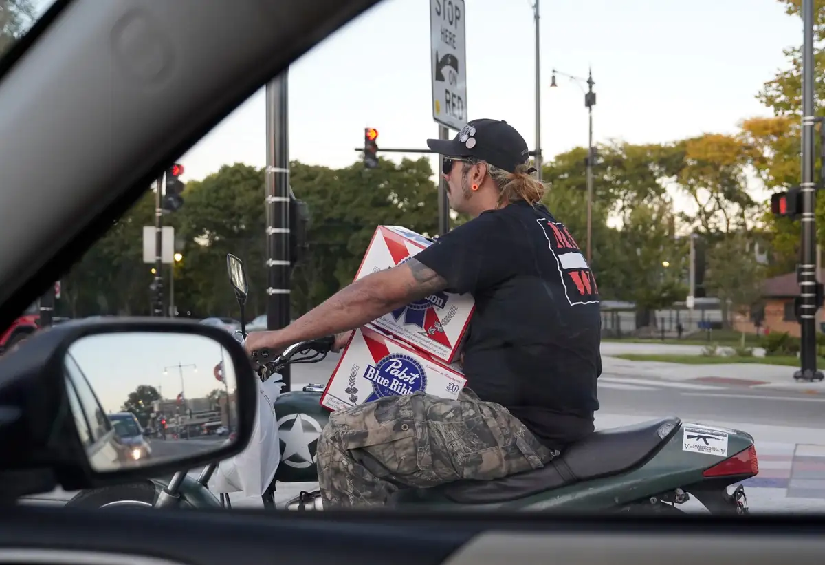 A man on a green motorcycle, viewed from inside a car, carries multiple large boxes of Pabst Blue Ribbon beer while stopped…